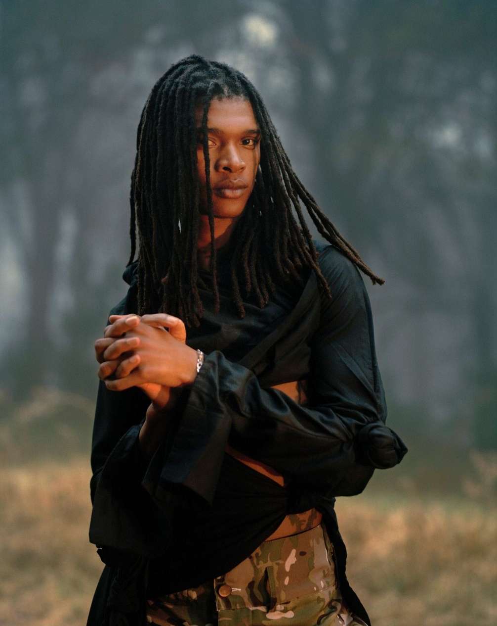 Person with dreadlocks wearing black shawl and camouflage pants in an outdoor setting with blurred trees in the background.
