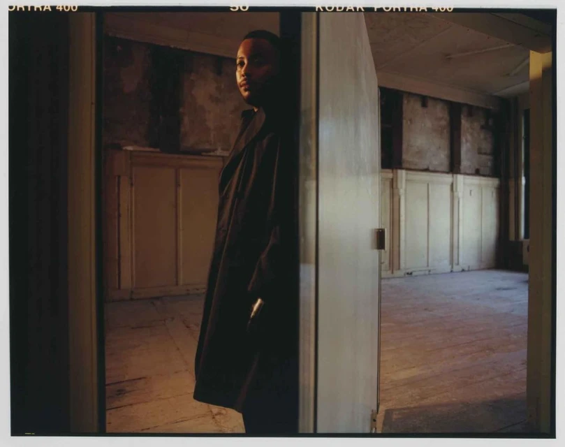 Indoor portrait of adult male wearing coat standing near doorway in room with pale walls and wooden floors with film border.