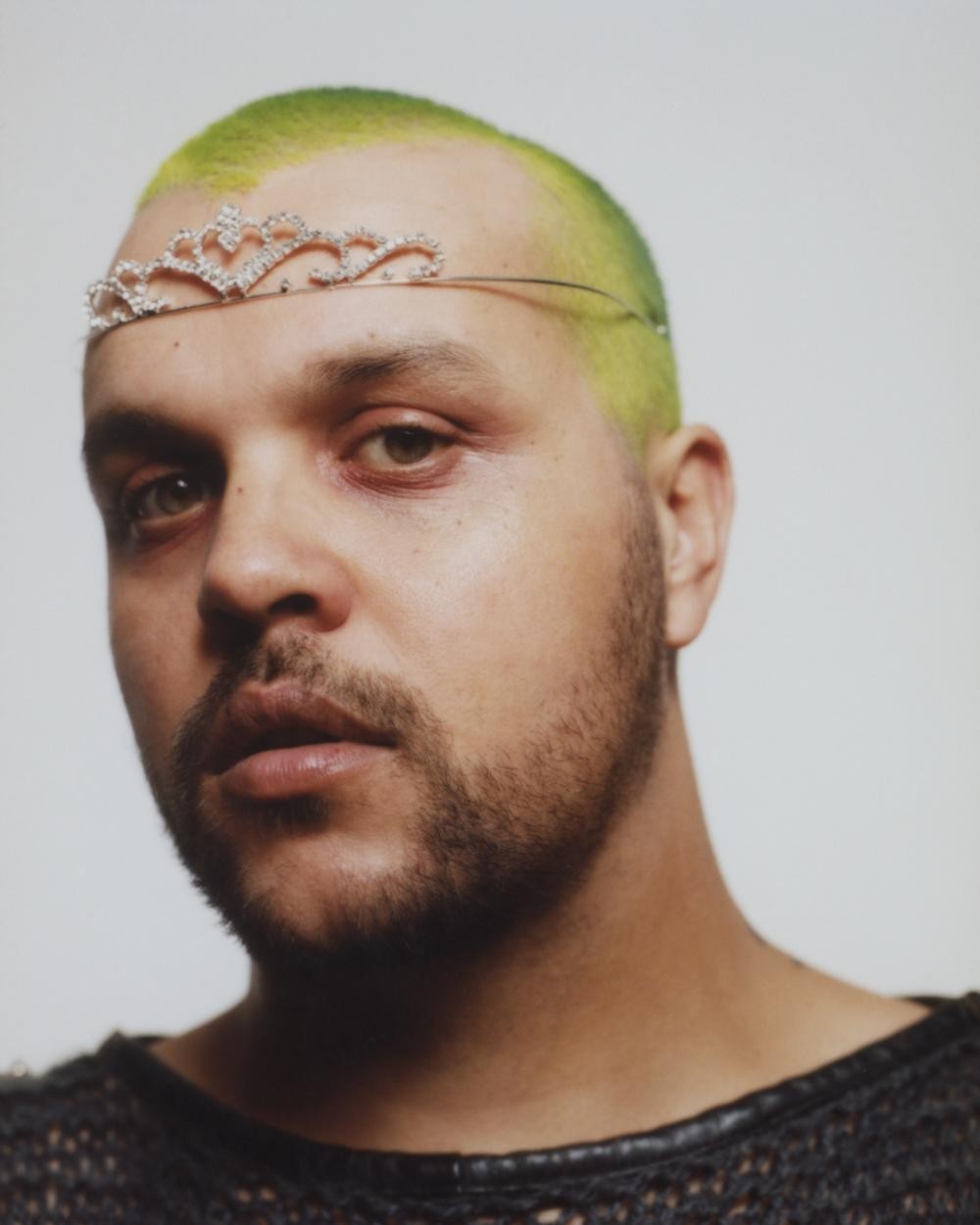 Close-up portrait of an adult man with green dyed hair and a decorative tiara, wearing a black knitted top, against a white backdrop.