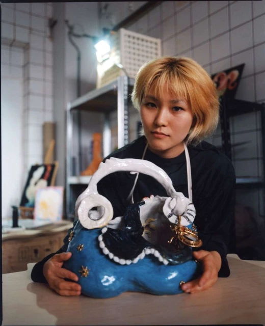 Woman with short blonde hair poses indoors while holding a blue, ceramic sculpture of a person wearing a black and white dress.
