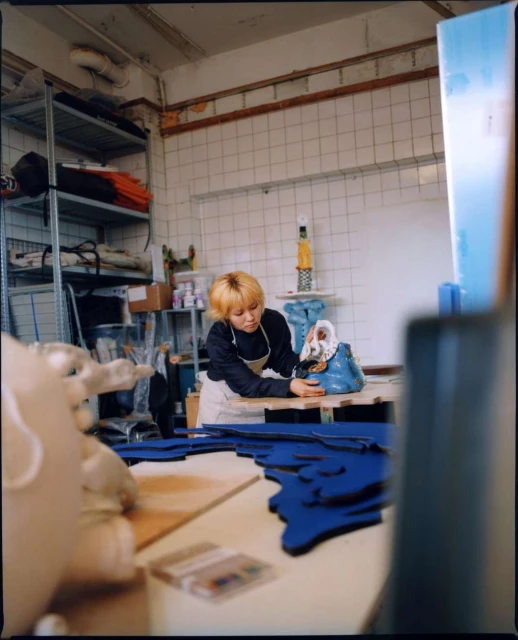 Young female artisan crafting a unique doll in a cluttered workshop with tools and blue fabric, in a bright indoor setting.