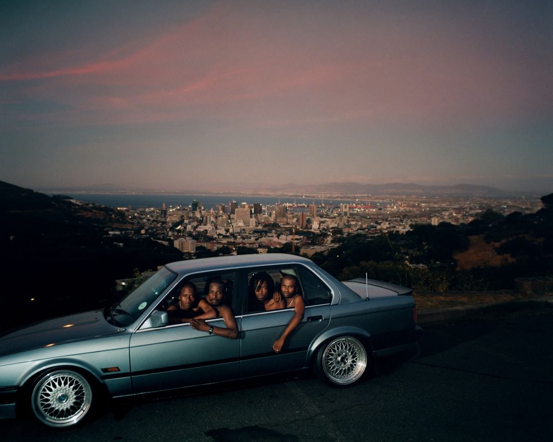 Four young adults in a vintage car overlooking a cityscape at dusk, with a pink and grey sky. Automotive and urban lifestyle photography.