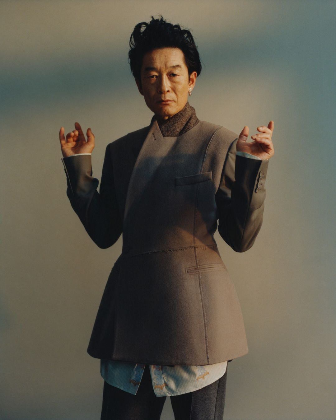Full-length portrait of an Asian adult male wearing a gray jacket with geometric tailoring, standing in front of a soft neutral background.