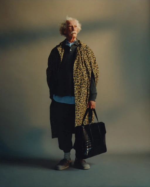 Full-length portrait of an elderly man with white hair wearing a leopard print coat, holding a black tote bag in a neutral studio.