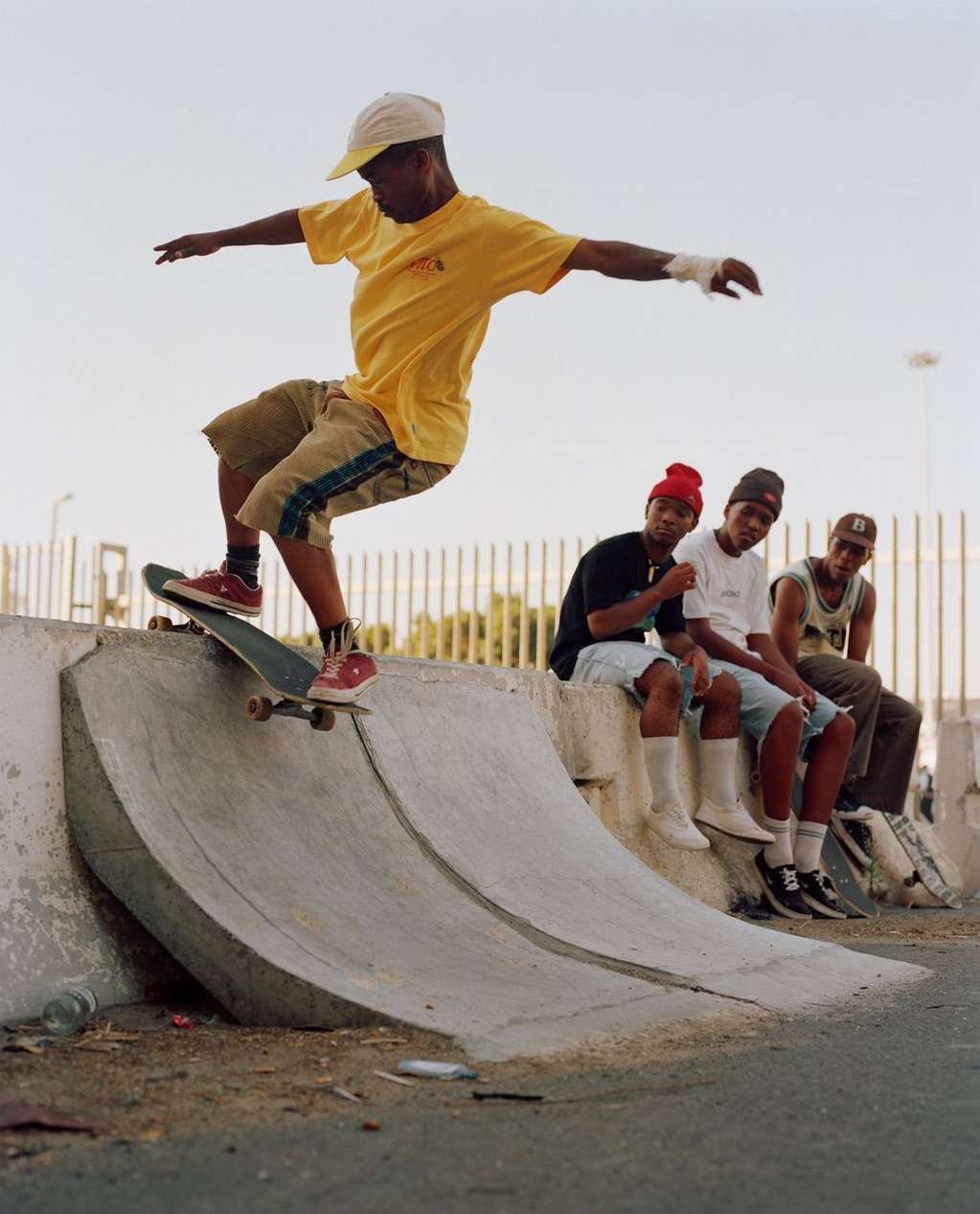 A skateboarder performs a trick on a concrete ramp while a group of young men sit and watch against a clear, bright sky in an outdoor setting.