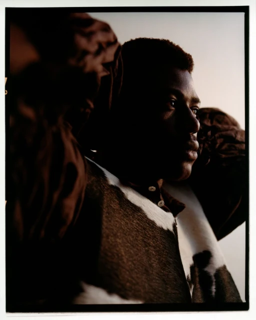 Portrait of a Black man in a patterned collared shirt. The image has a dark, shadowed aesthetic and a light-colored background.
