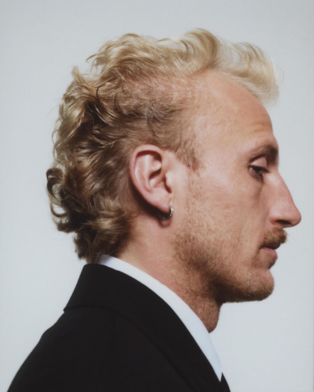 Profile portrait of an adult male with blond wavy hair and a mustache wearing a black suit jacket against a white background.
