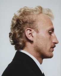 Profile portrait of a blonde man with a mustache wearing a black jacket and white collared shirt against a white background
