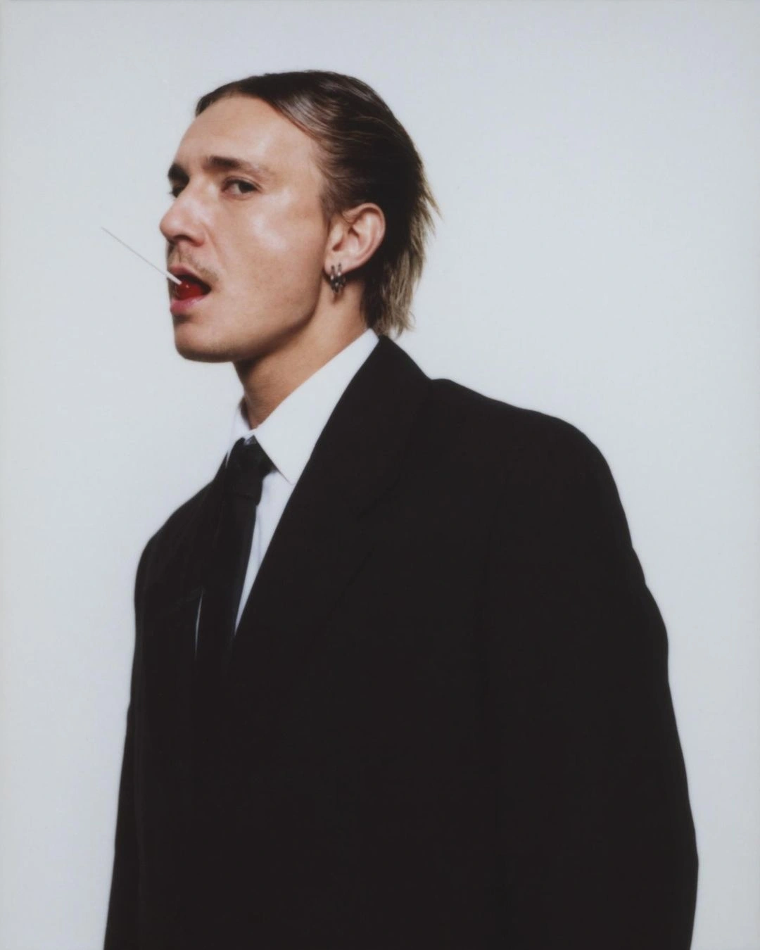 Portrait of a man in a black suit with a white shirt and black tie holding a lollipop in his mouth against a light grey background.