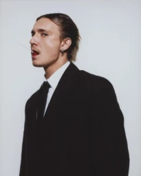 Portrait of a man in a black suit with a white shirt and black tie holding a lollipop in his mouth against a light grey background.