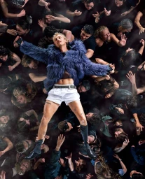A blond man crowdsurfing in a blue faux fur jacket and white shorts, supported by a dense crowd in a high-angle shot.