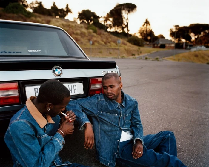 Two black men in denim jackets seated on asphalt leaning against a parked silver BMW car at dusk