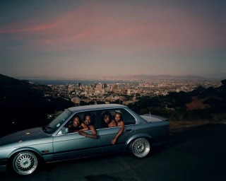Four Black men leaning out the windows of a silver sedan parked overlooking a city at dusk under a pink sky, by Nick Van Tiem