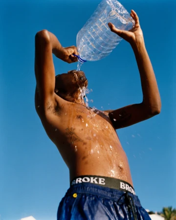 Shirtless man pouring a large plastic water bottle over his head against a clear blue sky background