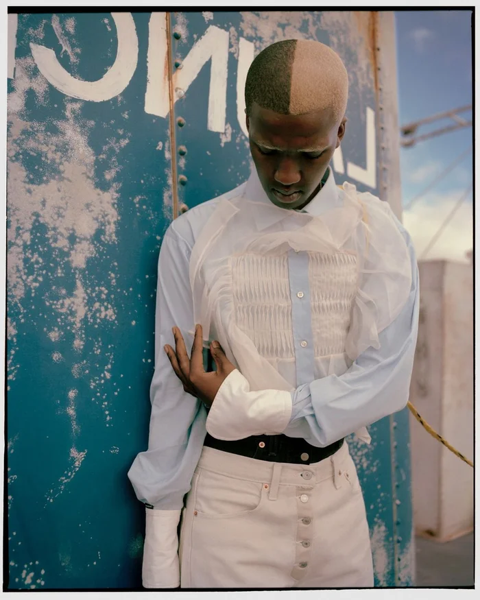 Portrait of a black man with dyed hair wearing a light blue shirt and white jeans against a weathered blue wall