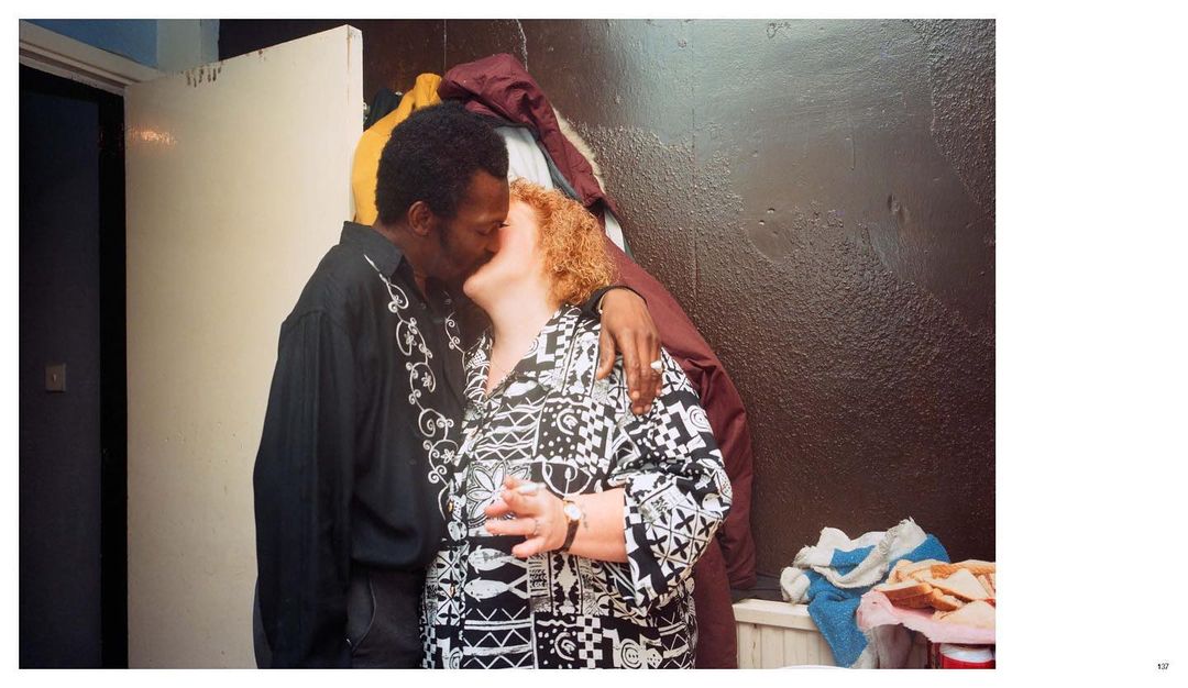 An adult African American male and a Caucasian female in a patterned shirt embrace indoors, near clothing and a painted dark wall.