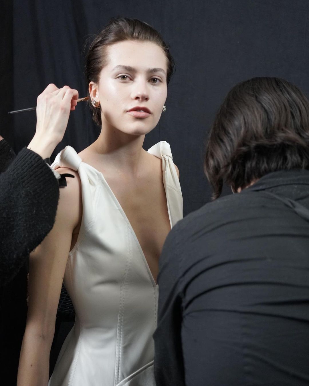 Woman in white dress getting makeup applied by makeup artist in studio with dark background during fashion or beauty photoshoot.