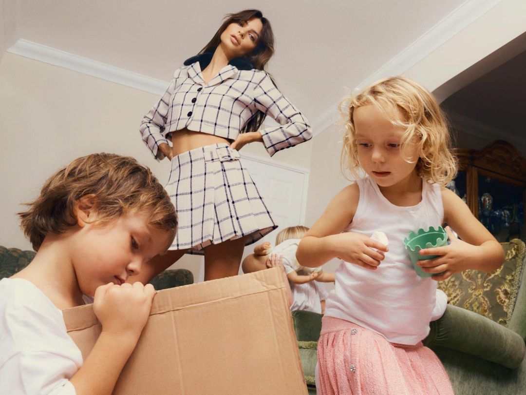 Fashion shot of a woman in a plaid skirt and crop top standing among four kids playing in a home interior with a white ceiling.