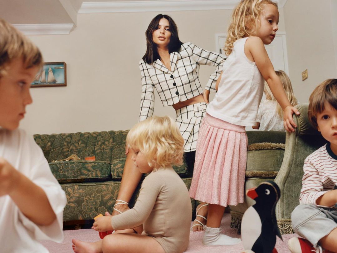 A woman in a plaid suit is surrounded by several children in an indoor setting, posing for a lifestyle fashion photo.