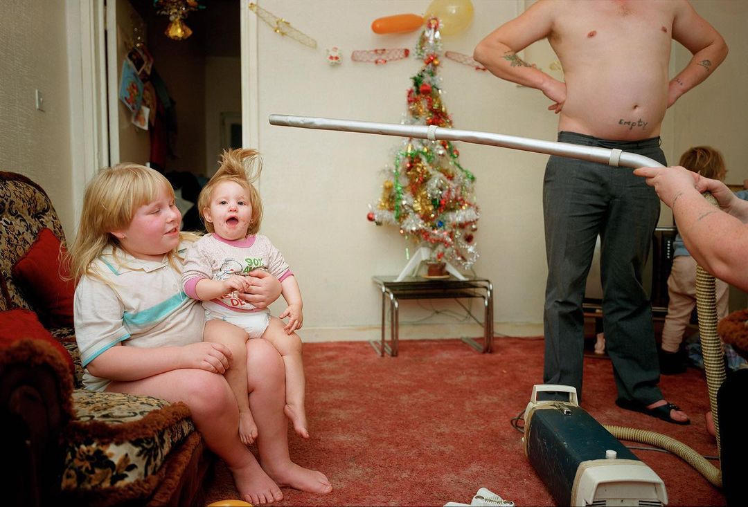 Candid family moment indoors featuring two kids and a man, with a vintage vacuum cleaner and Christmas decorations visible in the background.