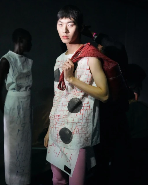 Runway fashion show featuring a male model wearing a printed tunic, leggings, and carrying a red mesh bag in a dark environment.