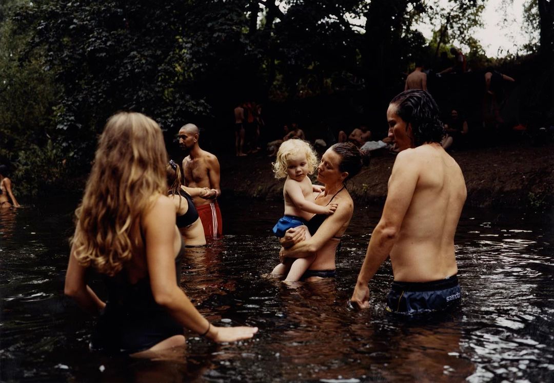 A group of adults and a child are cooling off in a flowing river surrounded by lush green foliage and trees on an overcast day.