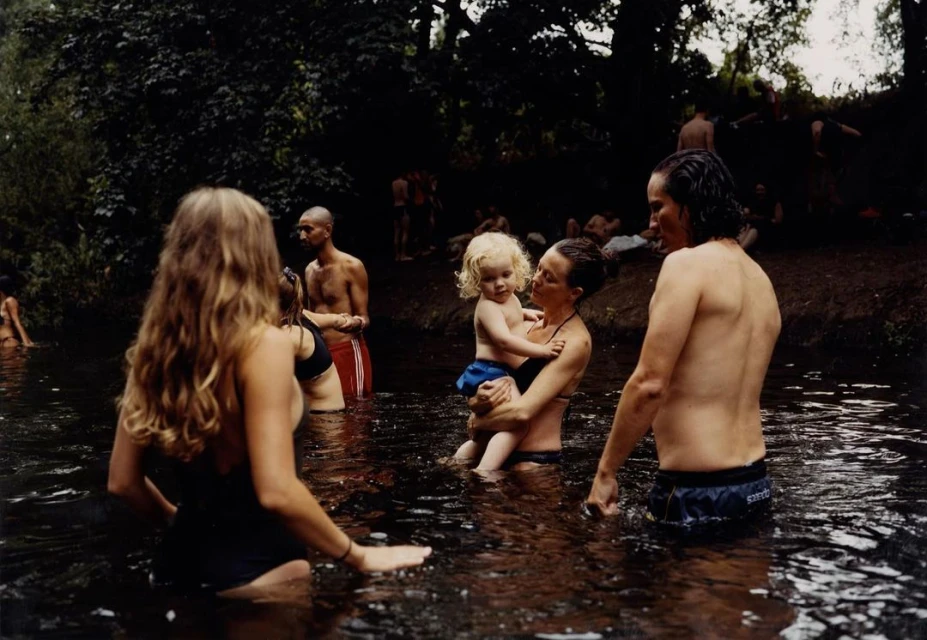 A group of adults and a child are cooling off in a flowing river surrounded by lush green foliage and trees on an overcast day.