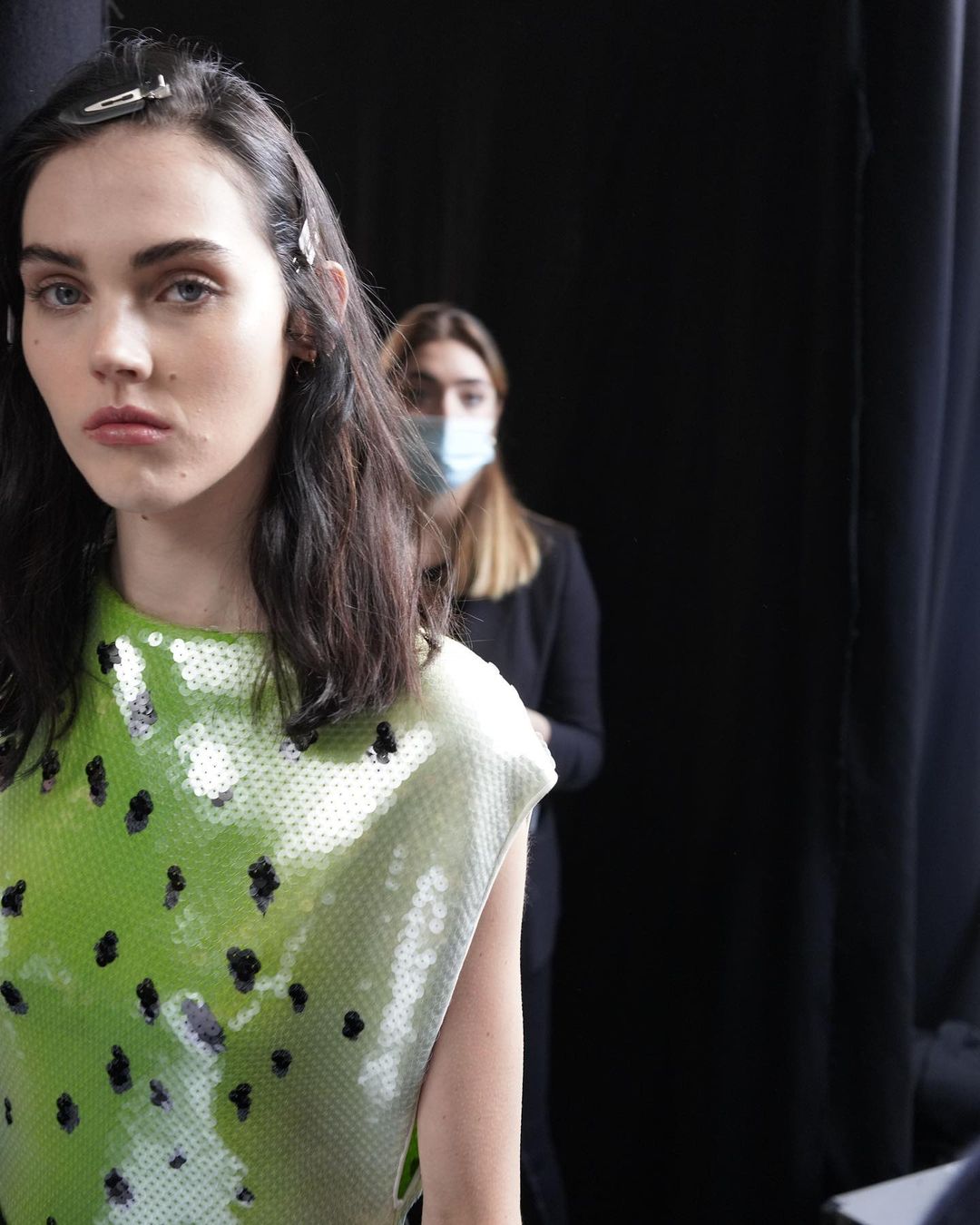 Backstage shot of a fashion model with brunette hair wearing a lime green and white sequined top, with a second woman in the background.