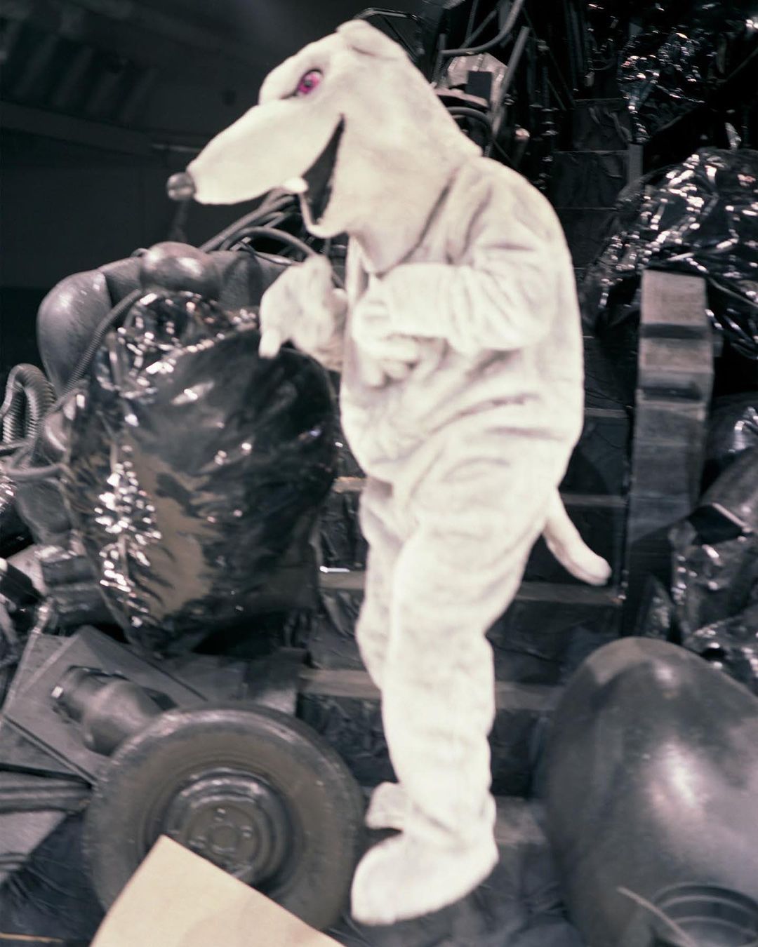 A person in a white animal costume stands among props and dark structures, illuminated by indoor lighting.