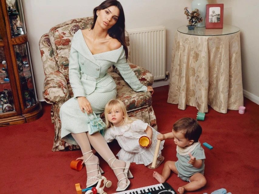 Model Emily Ratajkowski poses in a light-green skirt suit with two children in an indoor setting with floral upholstered furniture.
