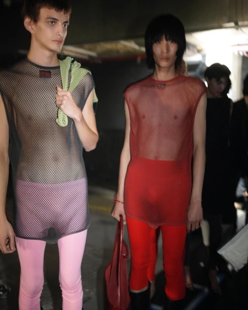 Two runway models backstage wearing sheer tops and leggings. One model holds a red tote bag in a dimly lit indoor environment.