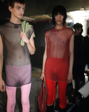 Two runway models backstage wearing sheer tops and leggings. One model holds a red tote bag in a dimly lit indoor environment.