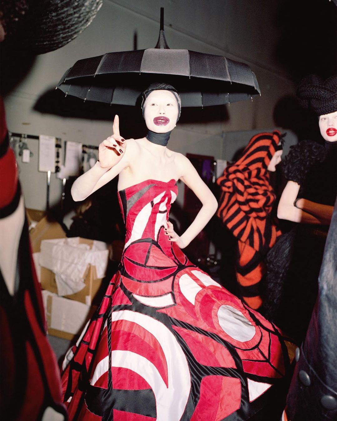 Avant-garde fashion shoot featuring a model in a red and white patterned dress, theatrical makeup, and an umbrella hat in an indoor setting.