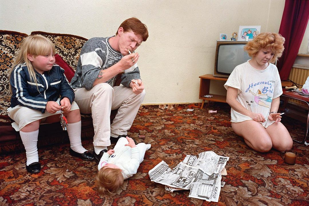 Family of four in a living room with patterned carpet and a television, two adults smoking cigarettes, a toddler on the floor, child on sofa.