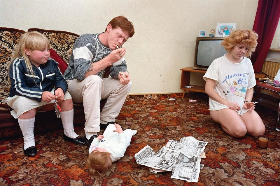 Family of four in a living room with patterned carpet and a television, two adults smoking cigarettes, a toddler on the floor, child on sofa.