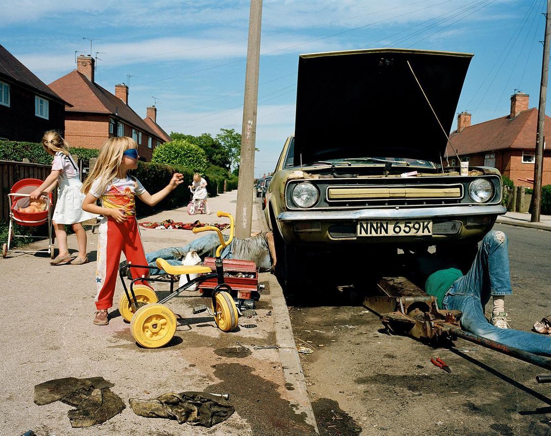 Residential street scene with children playing near a car being repaired, showcasing daily life in a suburban setting under bright daylight.