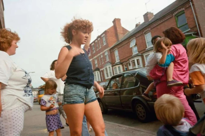 Outdoor scene showing an adult woman in denim shorts walking past a group with children on a street, next to parked cars.