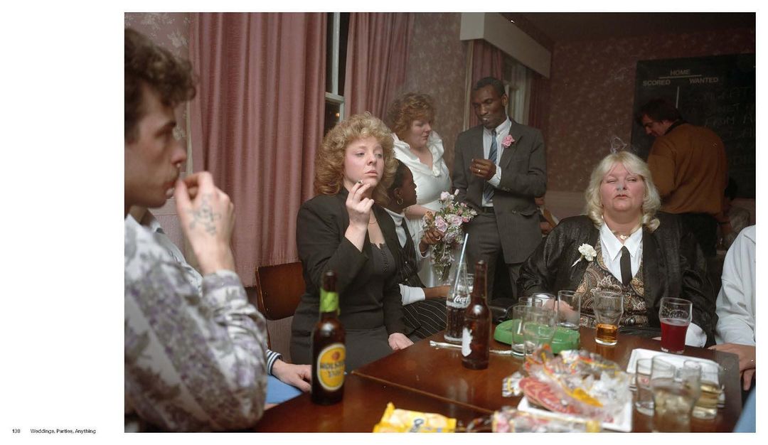 Indoor gathering of people around a table with drinks, with one woman smoking, and a man in a suit and tie visible in the background.