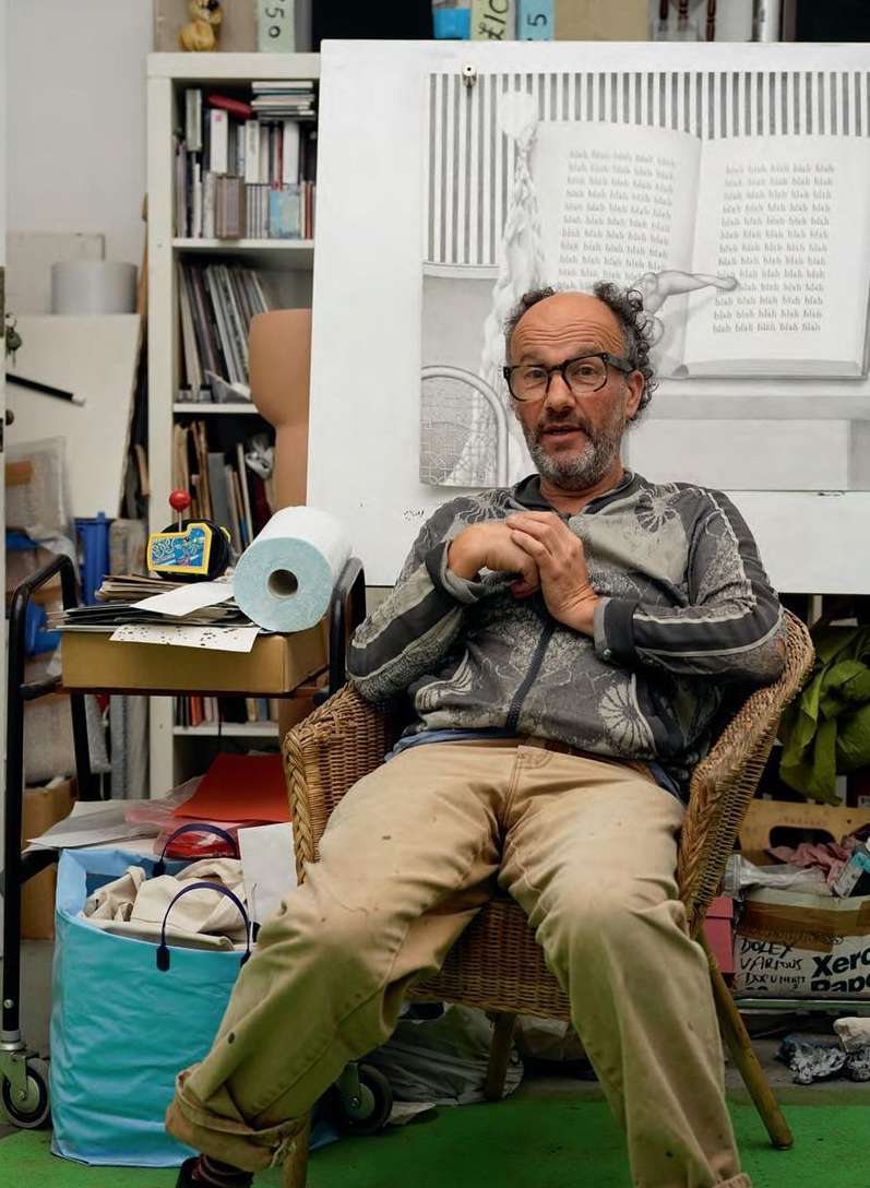 Man sitting in wicker chair in artist studio with large sketch behind him. Man is wearing glasses and patterned sweater.