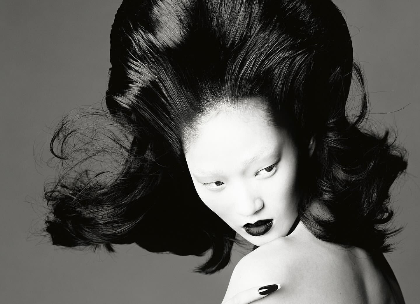 Monochrome portrait of Asian model with dramatic black hair and lipstick, neutral gray backdrop, beauty shot, high fashion editorial.