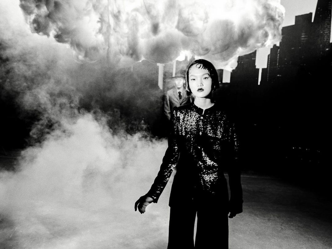 Monochrome portrait of an Asian woman in a sequined jacket and wide-leg pants, with a man in the background and cloud-like smoke.