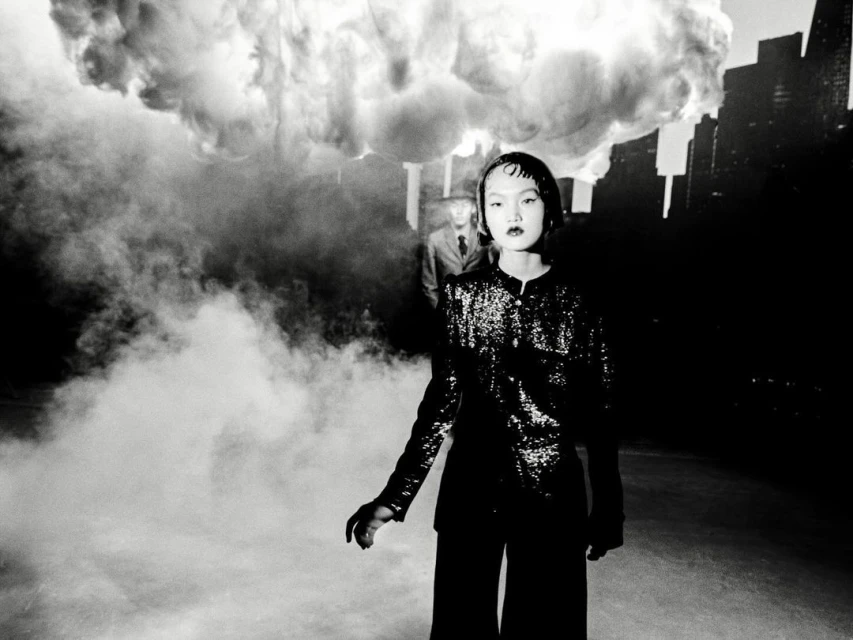 Monochrome portrait of an Asian woman in a sequined jacket and wide-leg pants, with a man in the background and cloud-like smoke.