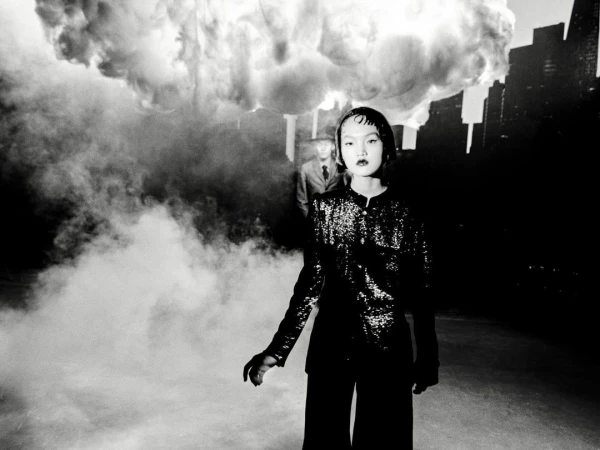 Monochrome portrait of an Asian woman in a sequined jacket and wide-leg pants, with a man in the background and cloud-like smoke.