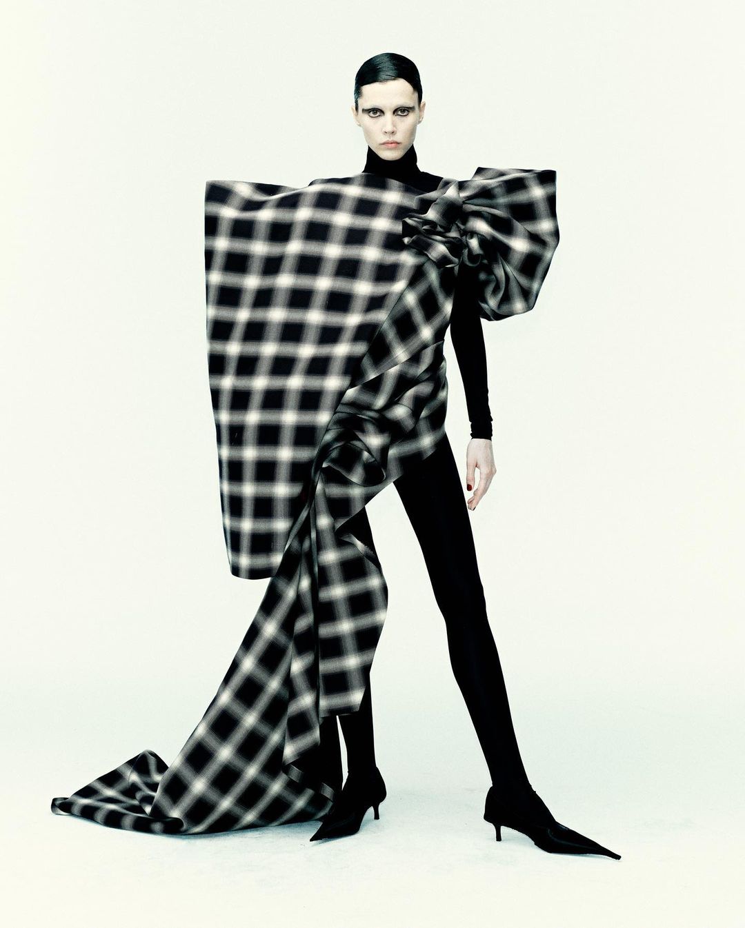 Fashion-forward model in black turtleneck and leggings with large plaid bow dress in a clean studio setting. Bold makeup and sleek hair.