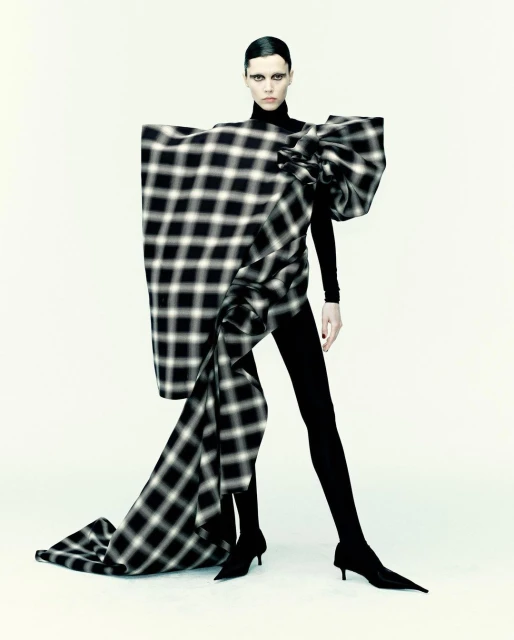 Fashion-forward model in black turtleneck and leggings with large plaid bow dress in a clean studio setting. Bold makeup and sleek hair.