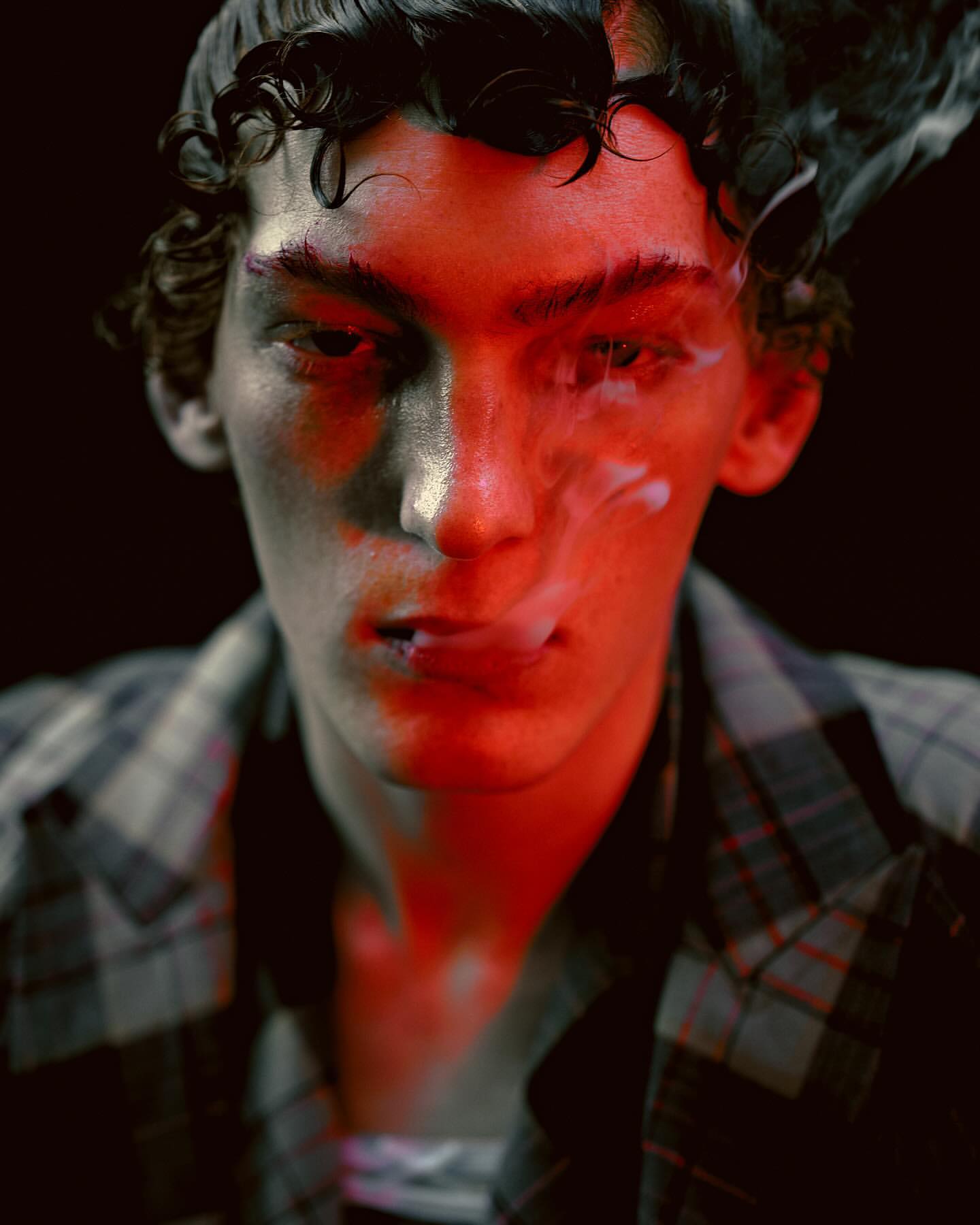 Close-up portrait of a young adult male with curly hair, wearing a plaid shirt, lit by red light, and exhaling white smoke against black.