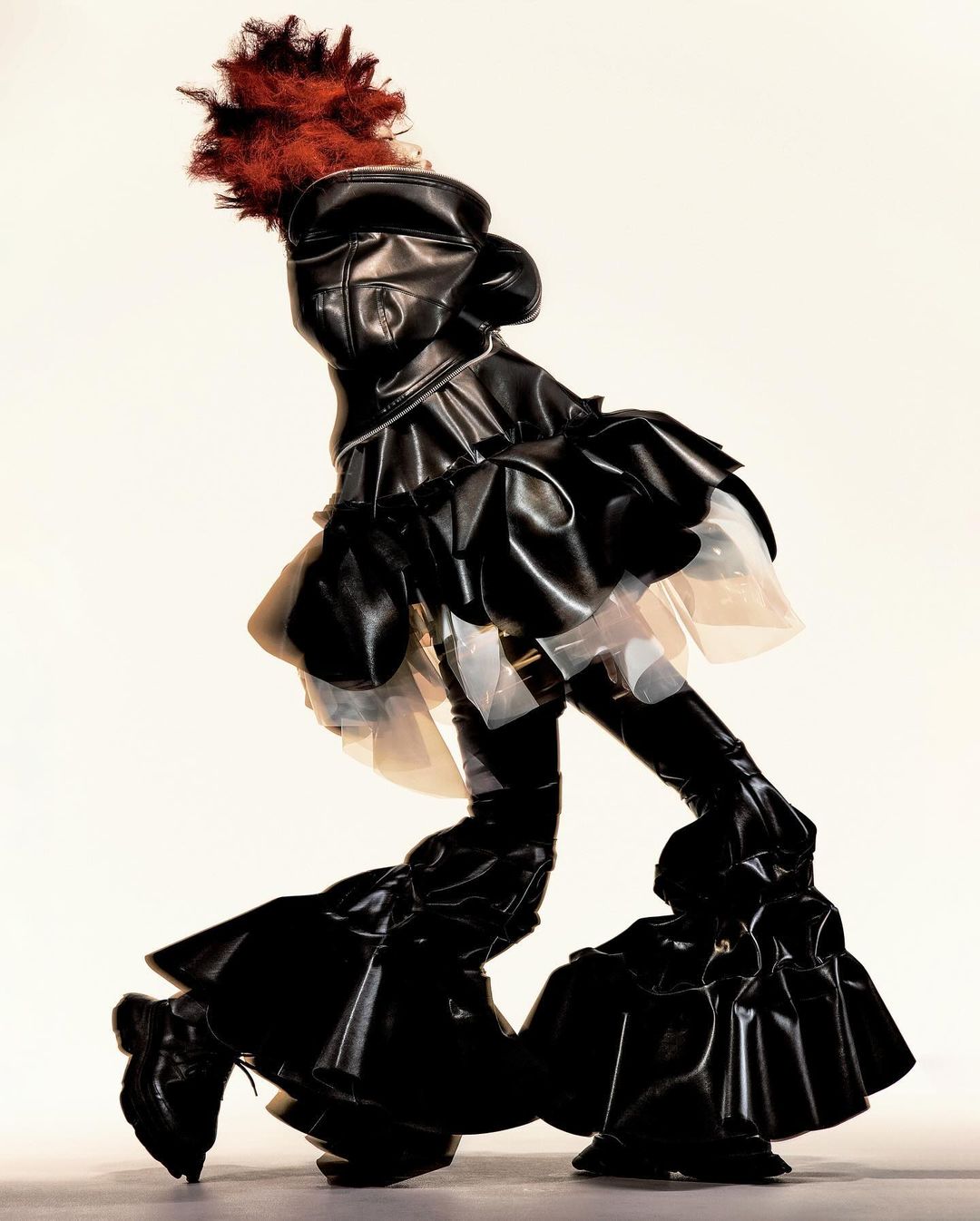 Person in black pleather ruffled avant-garde dress and shoes with red feather hat poses against a bright, neutral background in studio.