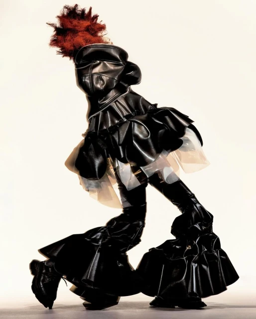 Person in black pleather ruffled avant-garde dress and shoes with red feather hat poses against a bright, neutral background in studio.