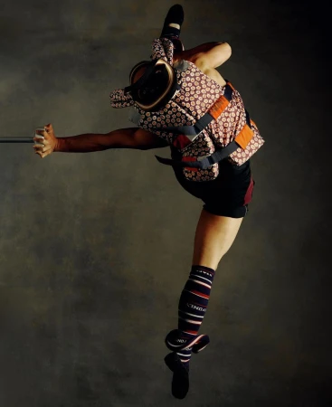 A lone adult is suspended mid-air in a ballet-style pose, showcasing unique patterned protective gear against a mottled grey backdrop.