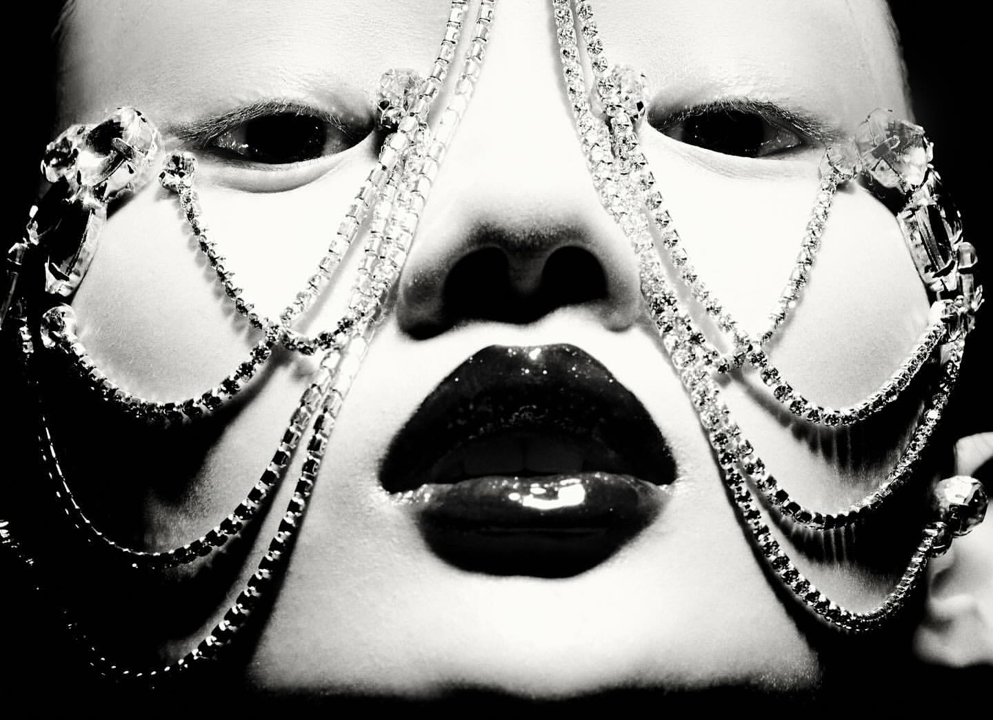 Close-up portrait of a face with a jeweled face covering, glossy lips, pale skin, and dark eyes, shot in black and white.